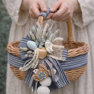 Puede incluir: Una cesta de mimbre tejida con una cinta y asa a rayas azules y blancas. La cesta está decorada con un lazo azul, flores secas, encaje y huevos decorativos. Un pequeño adorno con forma de pájaro y pompones blancos cuelgan de la cesta.