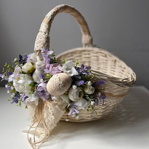 May include: A white wicker basket with a handle wrapped in lace. The basket is decorated with a bouquet of white and purple flowers, a small white bunny figurine, and a decorative egg.