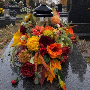 May include: A grave lantern with a black top and a small gold cross emblem, surrounded by an arrangement of artificial autumn-coloured flowers including deep red roses, bright yellow chrysanthemums, orange blossoms, and green foliage.