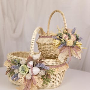 Può includere: Due cesti di vimini decorati per Pasqua. Il cesto più grande è di forma ovale e ha un manico. Il cesto più piccolo è rotondo e ha un manico. Entrambi i cesti sono decorati con pizzo, fiori e uova color pastello.