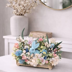 Könnte beinhalten: Dekorative Blumenanordnung in einer Holzkiste, mit einem hellblauen Hasen, pastellfarbenen Blumen und Schmetterlingsakzenten. Die Anordnung steht auf einem weißen Tisch, mit einer Vase mit weißen Blumen und einem Spiegel im Hintergrund.
