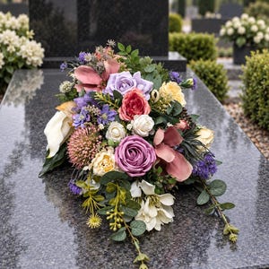 May include: A floral arrangement featuring a mix of artificial flowers in shades of purple, pink, yellow, and white, including roses, calla lilies, and spiky thistle-like blooms, accented with green eucalyptus leaves and stems on a polished stone surface.