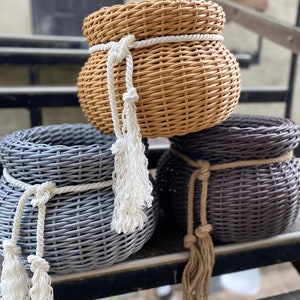 Puede incluir: Tres cestas tejidas en tonos beige, gris y marrón. Cada cesta está adornada con un detalle de cuerda alrededor de la parte superior y una borla a juego. Las cestas están dispuestas sobre una superficie oscura, mostrando sus texturas y colores.