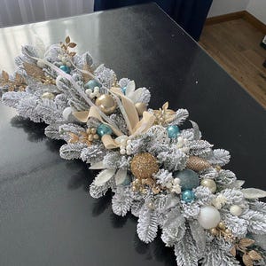 Puede incluir: Un festivo camino de mesa navideño con ramas de pino artificiales escarchadas, adornado con adornos dorados, plateados y verde azulado, cintas y detalles decorativos. El centro de mesa está dispuesto sobre una superficie oscura.