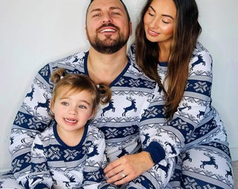 May include: Matching family pajamas in white and navy blue with a reindeer and snowflake pattern. The long-sleeved tops and pants are worn by a family of three. The pajamas have navy blue cuffs and trim.