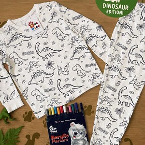 May include: White dinosaur-themed pajama set with long sleeves and pants, featuring black outline illustrations of various dinosaurs and the word "ROAR!". Includes a set of 12 acrylic markers for coloring. The set is labeled "Colour Me In Dinosaur Edition!"