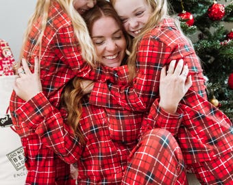 May include: Matching red, black, and white plaid button-down pajama sets featuring long sleeves and long pants. The fabric displays a classic tartan pattern with a collar, front button closure, and cuffs, worn by three individuals in a group.