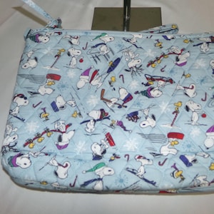 Op de afbeelding: Een lichtblauwe gewatteerde draagtas met een herhalend patroon van Snoopy-personages in wintersferen. De tas heeft een ritssluiting en een klein handvat. Het ontwerp bevat sneeuwvlokken en wintersport.