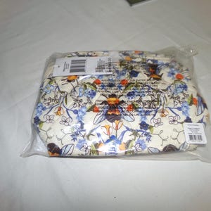 Puede incluir: Un neceser con estampado floral, empaquetado en una bolsa de plástico transparente. La bolsa presenta un fondo crema con motivos florales y de abeja azules, naranjas y marrones. Se adjunta una etiqueta con código de barras.
