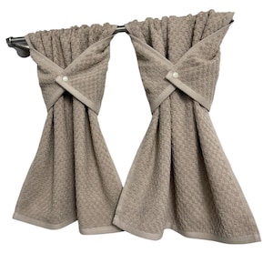 May include: Two beige kitchen towels hanging from a silver bar. The towels have a textured pattern and are folded with a decorative cross-over design, secured with a button. The towels have a light beige border.