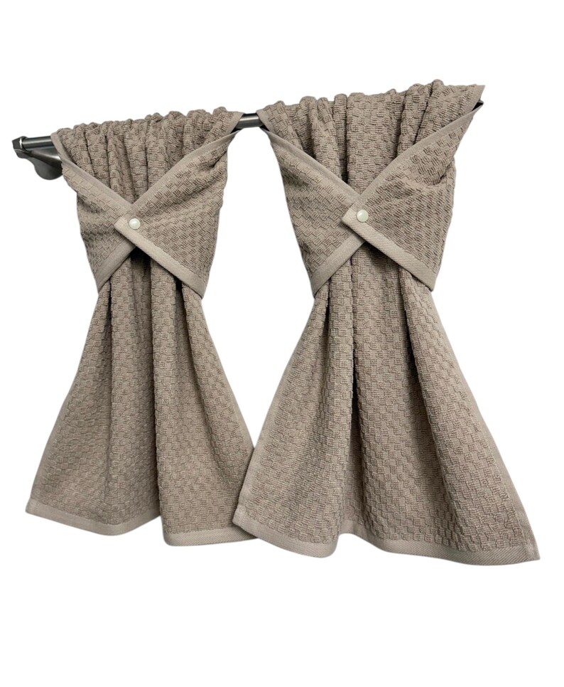 May include: Two beige kitchen towels hanging from a silver bar. The towels have a textured pattern and are folded with a decorative cross-over design, secured with a button. The towels have a light beige border.