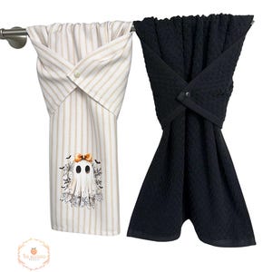 May include: Two decorative kitchen towels hanging on a silver bar. One towel is white with vertical gold stripes and a ghost graphic. The other towel is solid black. Both towels are folded and secured with a button.