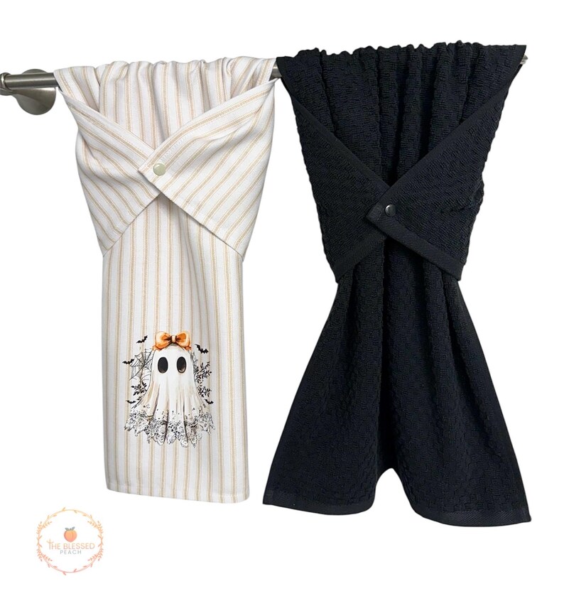 May include: Two decorative kitchen towels hanging on a silver bar. One towel is white with vertical gold stripes and a ghost graphic. The other towel is solid black. Both towels are folded and secured with a button.
