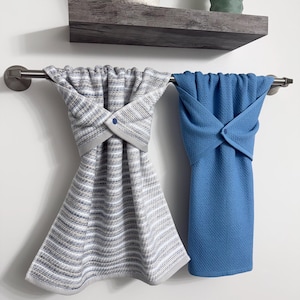 May include: Two decorative hand towels hanging on a metal bar. One towel is blue, and the other is gray and white striped. Both towels are folded with a button closure. A shelf with a plant and candle is above the towels.