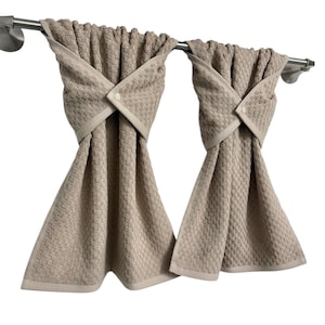 May include: Two beige kitchen towels with a textured pattern are hanging on a stainless steel bar. Each towel is folded and secured with a button closure. The towels have a light beige border.