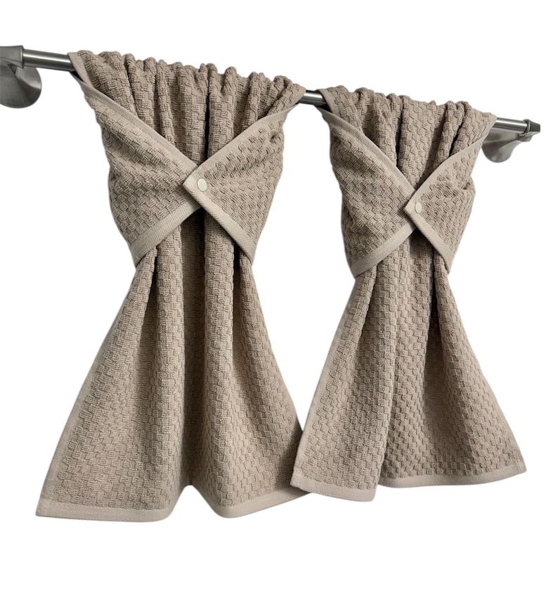 May include: Two beige kitchen towels with a textured pattern are hanging on a stainless steel bar. Each towel is folded and secured with a button closure. The towels have a light beige border.