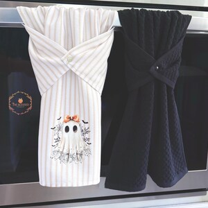 May include: Two decorative kitchen towels. One is white with vertical tan stripes and a ghost graphic with a bow. The other is solid black with a textured pattern. Both are looped over a bar and secured with a button.