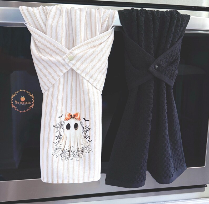 May include: Two decorative kitchen towels. One is white with vertical tan stripes and a ghost graphic with a bow. The other is solid black with a textured pattern. Both are looped over a bar and secured with a button.