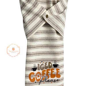One stay put kitchen towel with brown stripes on an ivory background. The towel has the text 'Iced Coffee Please' printed on the front of the towel in orange and brown lettering with a black coffee cup on each side of the text. The towel has a snap closure so it can stay in place on an oven/stove/dishwasher handle. This listing is for one towel and includes free shipping.