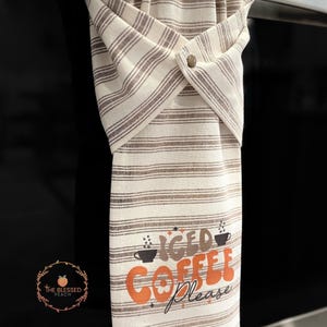 One stay put kitchen towel with brown stripes on an ivory background. The towel has the text 'Iced Coffee Please' printed on the front of the towel in orange and brown lettering with a black coffee cup on each side of the text. The towel has a snap closure so it can stay in place on an oven/stove/dishwasher handle. This listing is for one towel and includes free shipping.