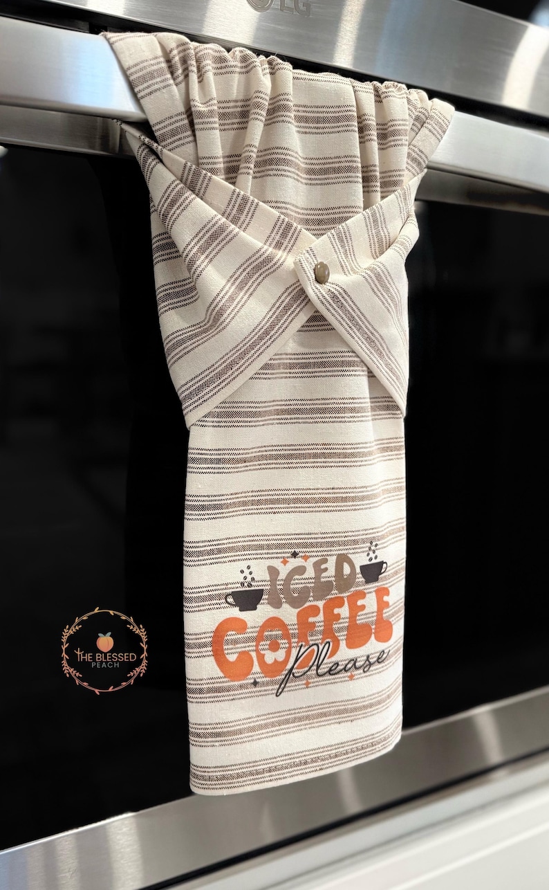 One stay put kitchen towel with brown stripes on an ivory background. The towel has the text 'Iced Coffee Please' printed on the front of the towel in orange and brown lettering with a black coffee cup on each side of the text. The towel has a snap closure so it can stay in place on an oven/stove/dishwasher handle. This listing is for one towel and includes free shipping.