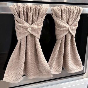 May include: Two beige kitchen towels hanging on an oven handle. The towels are folded and secured with a button closure. The towels have a textured weave pattern and a light beige trim.
