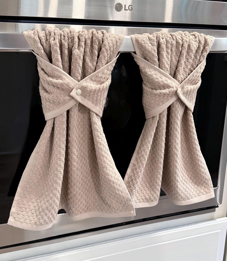 May include: Two beige kitchen towels hanging on an oven handle. The towels are folded and secured with a button closure. The towels have a textured weave pattern and a light beige trim.