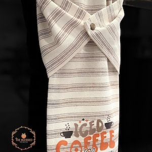 One stay put kitchen towel with brown stripes on an ivory background. The towel has the text 'Iced Coffee Please' printed on the front of the towel in orange and brown lettering with a black coffee cup on each side of the text. The towel has a snap closure so it can stay in place on an oven/stove/dishwasher handle. This listing is for one towel and includes free shipping.
