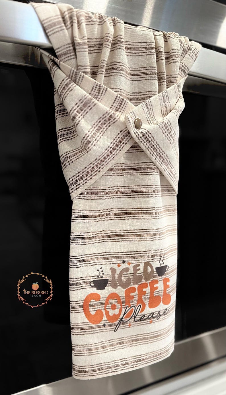 One stay put kitchen towel with brown stripes on an ivory background. The towel has the text 'Iced Coffee Please' printed on the front of the towel in orange and brown lettering with a black coffee cup on each side of the text. The towel has a snap closure so it can stay in place on an oven/stove/dishwasher handle. This listing is for one towel and includes free shipping.