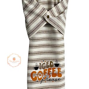 One stay put kitchen towel with brown stripes on an ivory background. The towel has the text 'Iced Coffee Please' printed on the front of the towel in orange and brown lettering with a black coffee cup on each side of the text. The towel has a snap closure so it can stay in place on an oven/stove/dishwasher handle. This listing is for one towel and includes free shipping.