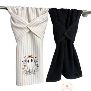 May include: Two Halloween-themed kitchen towels hanging on a metal bar. One towel is white with tan stripes and a ghost graphic. The other is solid black. Both towels are folded and buttoned at the top.