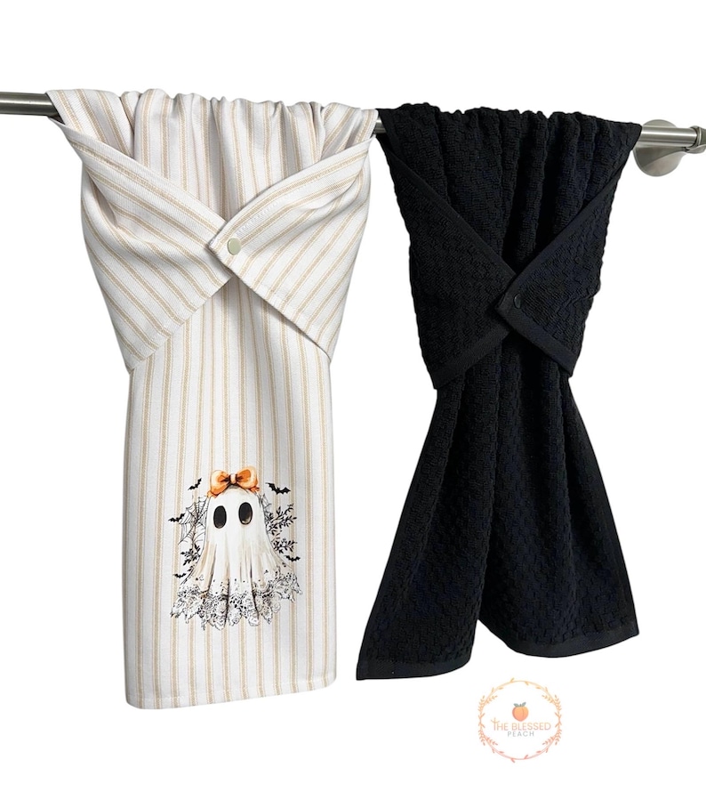 May include: Two Halloween-themed kitchen towels hanging on a metal bar. One towel is white with tan stripes and a ghost graphic. The other is solid black. Both towels are folded and buttoned at the top.