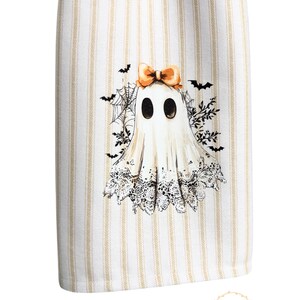 May include: White and beige striped kitchen towel featuring a Halloween ghost design. The ghost has a bow, lace trim, and is surrounded by bats and spiderwebs. The towel has a decorative design.