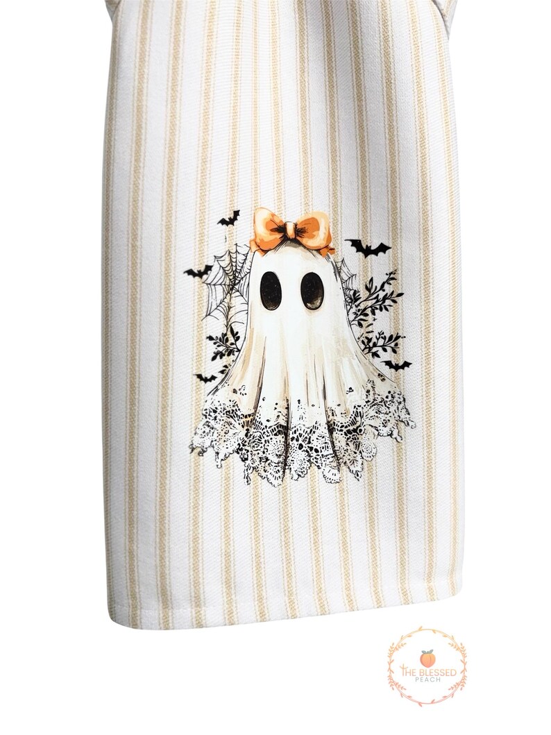 May include: White and beige striped kitchen towel featuring a Halloween ghost design. The ghost has a bow, lace trim, and is surrounded by bats and spiderwebs. The towel has a decorative design.