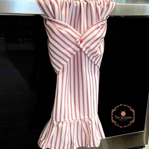 May include: A red and white striped kitchen towel with a ruffled edge hangs on a silver oven handle. The towel has a single red button on the front. The text "The Blessed Peach" is visible on the oven door.