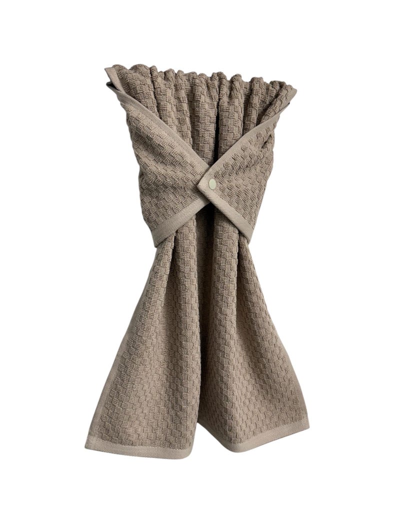 May include: A beige kitchen towel with a woven pattern. The towel is folded and secured with a button closure. The towel has a light beige border and is designed for hanging.