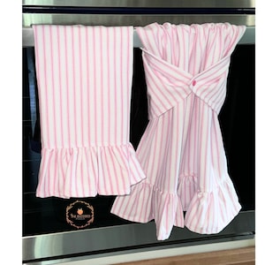 May include: Two pink and white striped kitchen towels with ruffled edges. One towel hangs straight, while the other is folded and buttoned. The towels are displayed on a stainless steel oven.