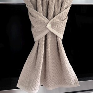 May include: A beige kitchen towel with a textured weave, hanging from a stainless steel appliance. The towel is folded and secured with a button, creating a decorative and functional design for the kitchen.