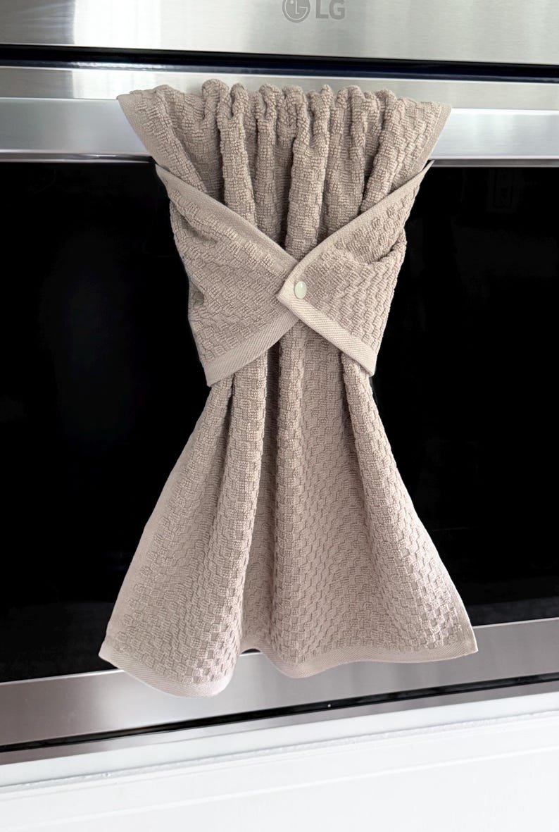 May include: A beige kitchen towel with a textured weave, hanging from a stainless steel appliance. The towel is folded and secured with a button, creating a decorative and functional design for the kitchen.