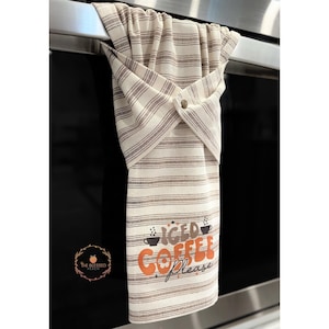 One stay put kitchen towel with brown stripes on an ivory background. The towel has the text 'Iced Coffee Please' printed on the front of the towel in orange and brown lettering with a black coffee cup on each side of the text. The towel has a snap closure so it can stay in place on an oven/stove/dishwasher handle. This listing is for one towel and includes free shipping.