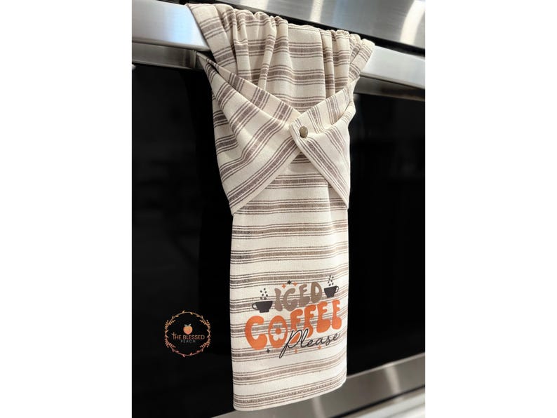 One stay put kitchen towel with brown stripes on an ivory background. The towel has the text 'Iced Coffee Please' printed on the front of the towel in orange and brown lettering with a black coffee cup on each side of the text. The towel has a snap closure so it can stay in place on an oven/stove/dishwasher handle. This listing is for one towel and includes free shipping.
