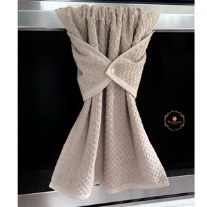 May include: A beige kitchen towel with a textured weave, hanging from a metal bar. The towel is folded and secured with a button closure. The Blessed Peach logo is visible on the right side.