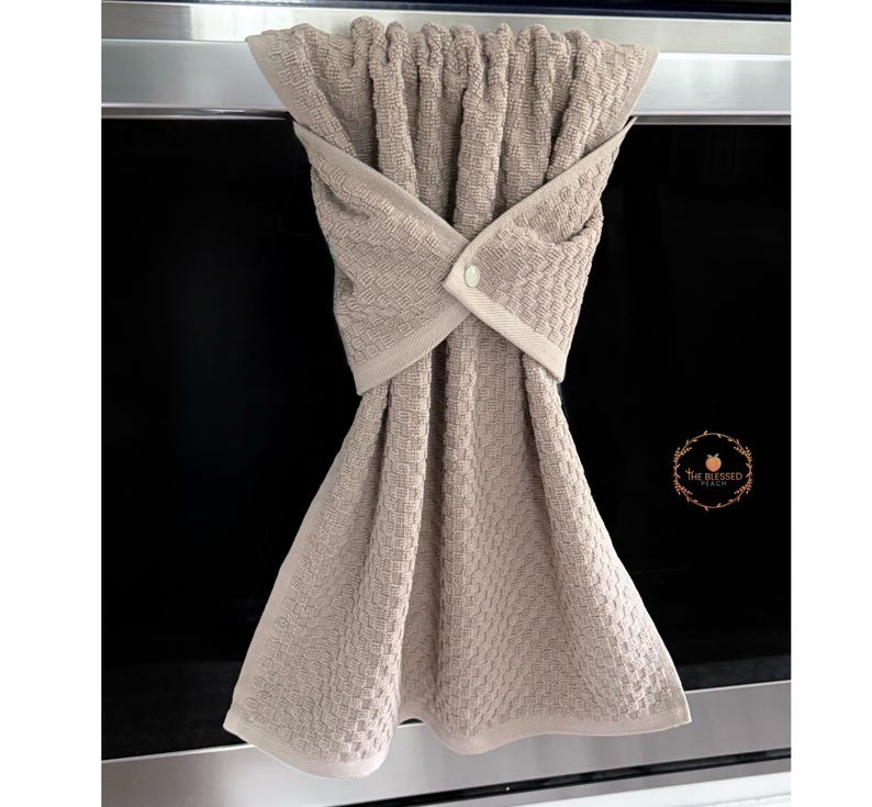May include: A beige kitchen towel with a textured weave, hanging from a metal bar. The towel is folded and secured with a button closure. The Blessed Peach logo is visible on the right side.