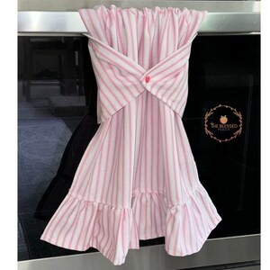 May include: A pink and white striped kitchen towel with a ruffled bottom and a button closure. The towel is hanging on an oven handle. The Blessed Peach logo is visible on the oven.