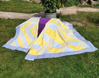 May include: A yellow and blue patchwork quilt with a floral pattern. The quilt is folded and laid out on a grassy surface.