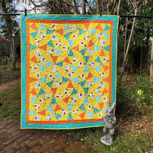 May include: A yellow, orange and blue quilt with a geometric pattern. The quilt is hanging on a clothesline in a garden setting.