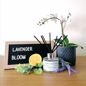 May include: A clear glass bottle with a black lid and black reeds, filled with a lavender-scented reed diffuser. The bottle has a label that reads "The Surrey Hills Candle Co. Lavender Bloom Reed Diffuser". The bottle is surrounded by a lemon, eucalyptus leaves, and lavender flowers.