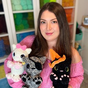 May include: A woman is holding three crocheted cat toys. One is white with a pink bow, one is gray with a black eye patch, and one is black with an orange witch hat.
