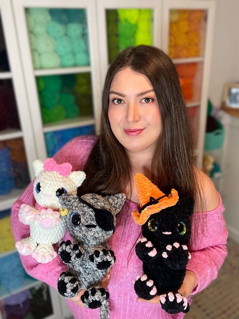May include: A woman is holding three crocheted cat toys. One is white with a pink bow, one is gray with a black eye patch, and one is black with an orange witch hat.