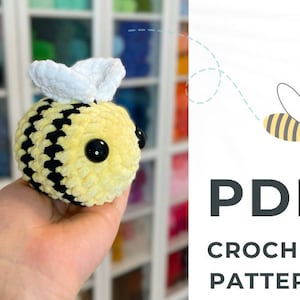 May include: A crocheted bee amigurumi toy with yellow and black stripes, white wings, and black eyes. The bee is being held in a hand. The image includes the text "PDF CROCHET PATTERN".